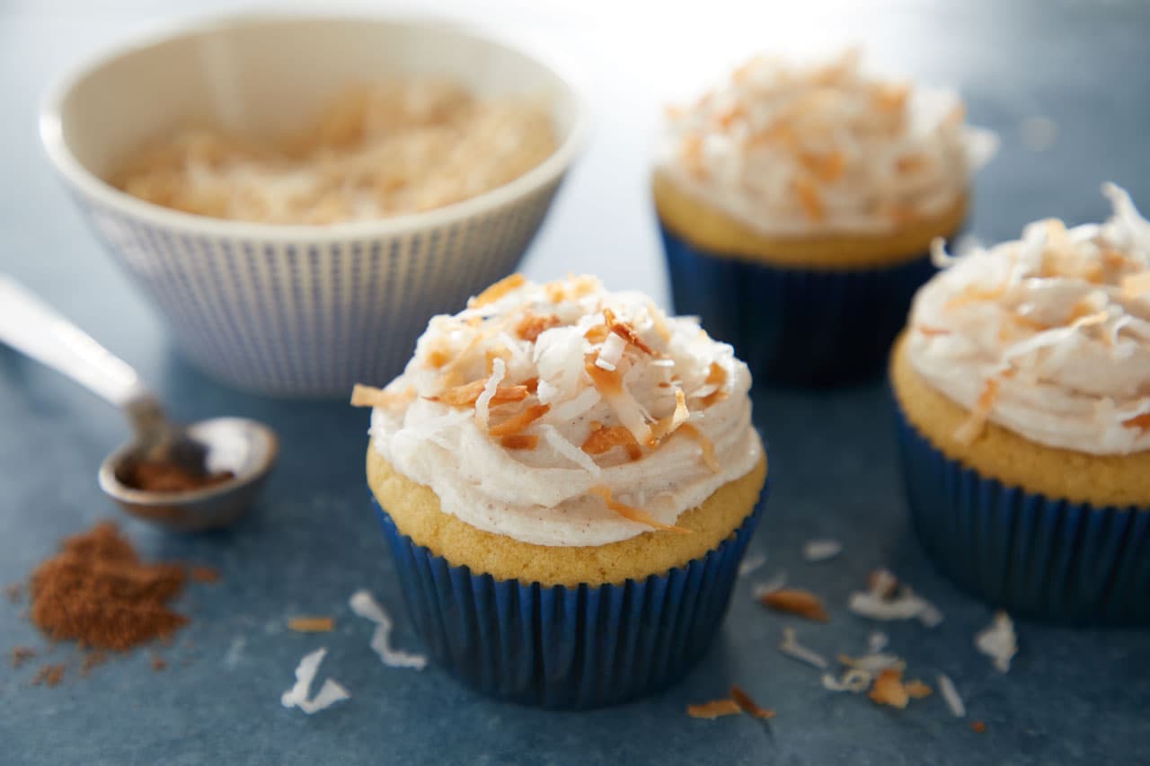 Three cupcakes with white frosting and toasted coconut topping are arranged on a blue surface, with a bowl of shredded coconut and a spoon with spice in the background.