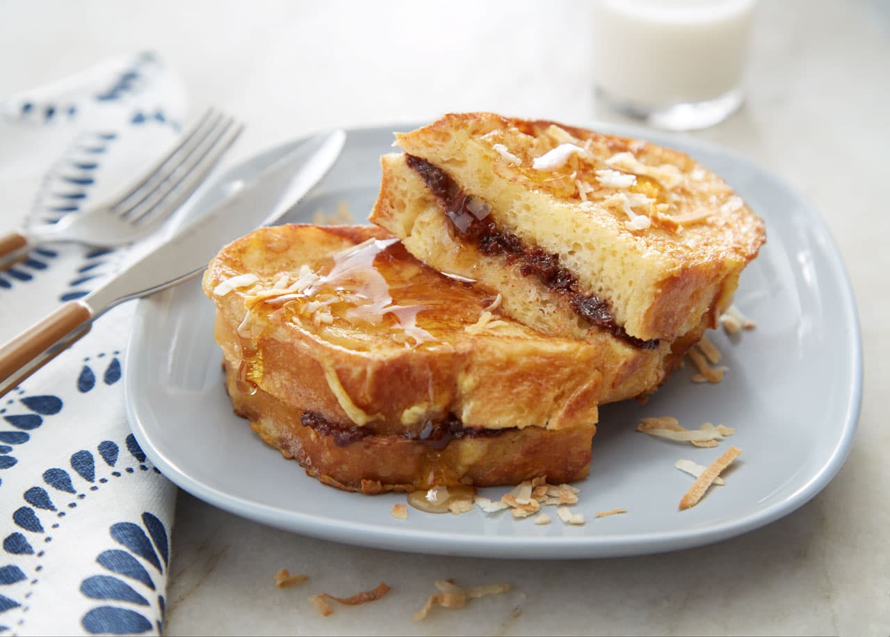 Two slices of stuffed French toast with a chocolate filling, topped with toasted coconut, served on a gray plate with a fork, knife, and a glass of milk in the background.