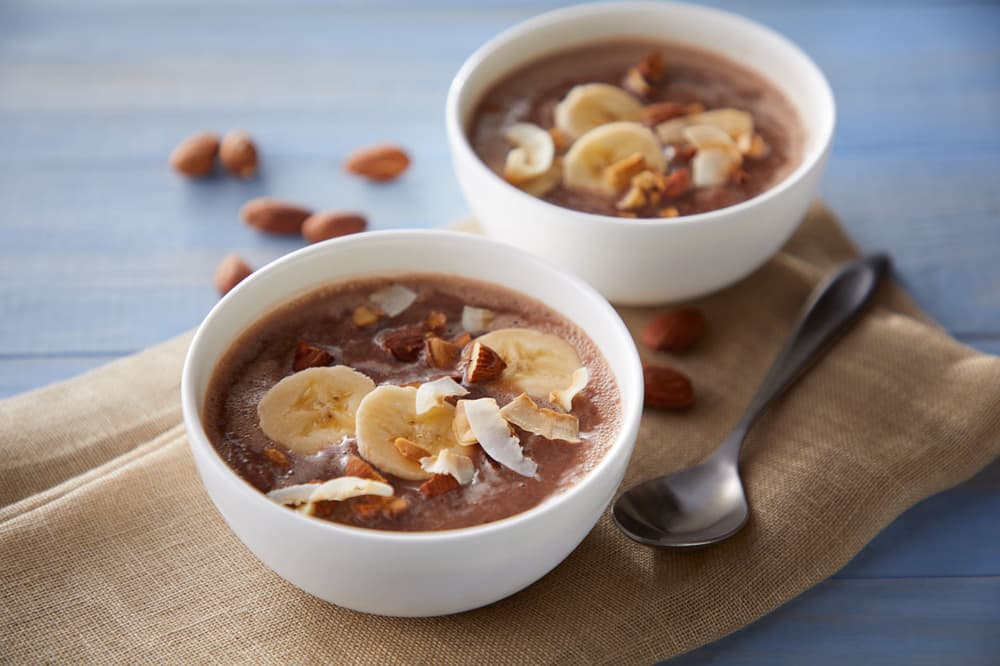Two white bowls of smoothie topped with banana slices, nuts, and coconut flakes on a wooden surface with a spoon and scattered almonds nearby.