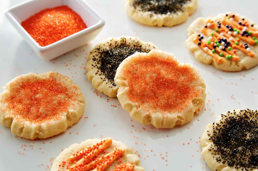 Assorted sugar cookies decorated with orange and black sprinkles are arranged on a white surface next to a small square bowl of orange sanding sugar.