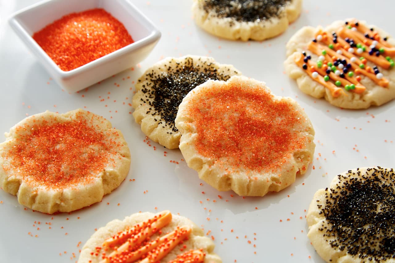 Assorted sugar cookies decorated with orange and black sprinkles are arranged on a white surface next to a small square bowl of orange sanding sugar.