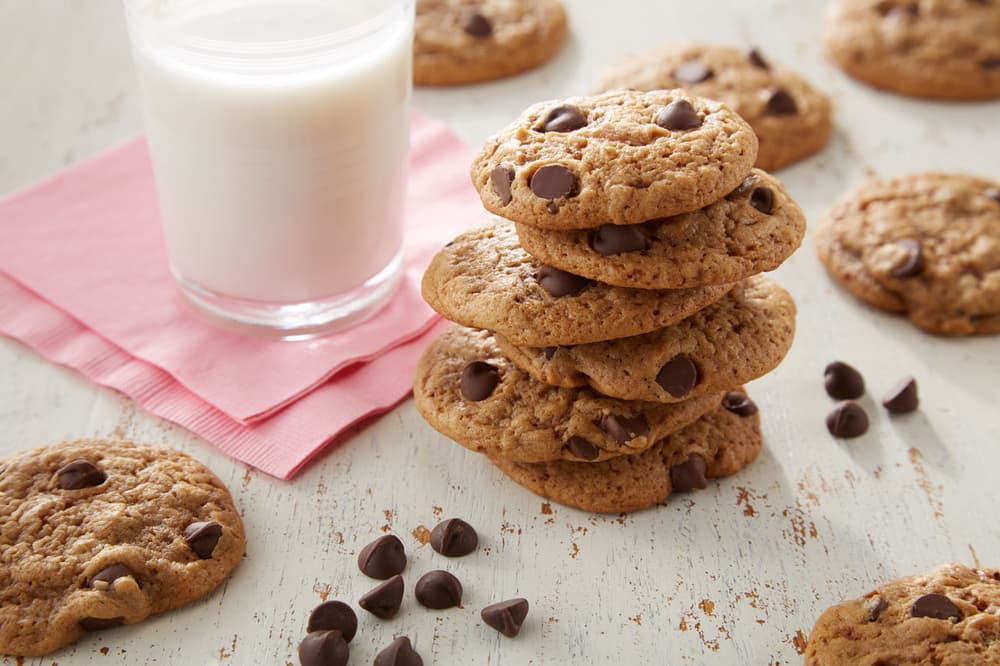 A stack of chocolate chip cookies sits next to a glass of milk on a pink napkin, with more cookies and chocolate chips scattered on a white wooden surface.