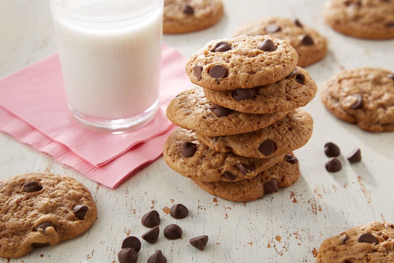 A stack of chocolate chip cookies sits next to a glass of milk on a pink napkin, with more cookies and chocolate chips scattered on a white wooden surface.