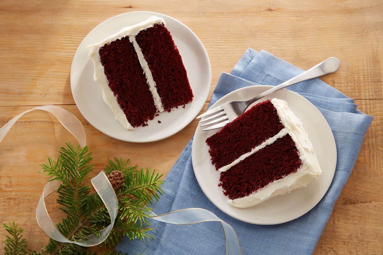 Two slices of red velvet cake with white frosting on white plates, one plate on a blue napkin with a fork, next to a pine branch and ribbon on a wooden surface.