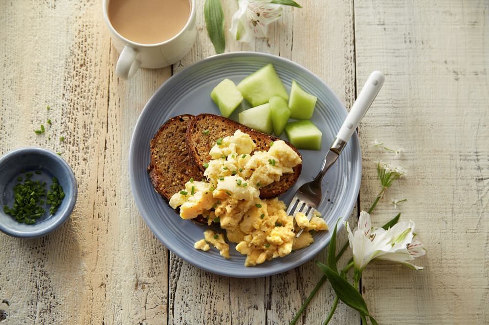 A plate with scrambled eggs, two slices of toast, and honeydew melon cubes, next to a cup of coffee and a small bowl of chopped chives on a rustic wooden table.