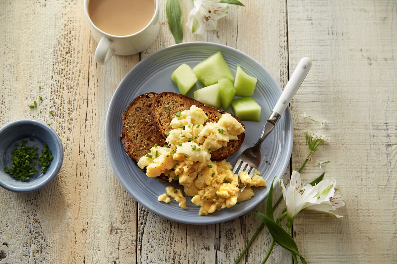 A plate with scrambled eggs, two slices of toast, and honeydew melon cubes, next to a cup of coffee and a small bowl of chopped chives on a rustic wooden table.