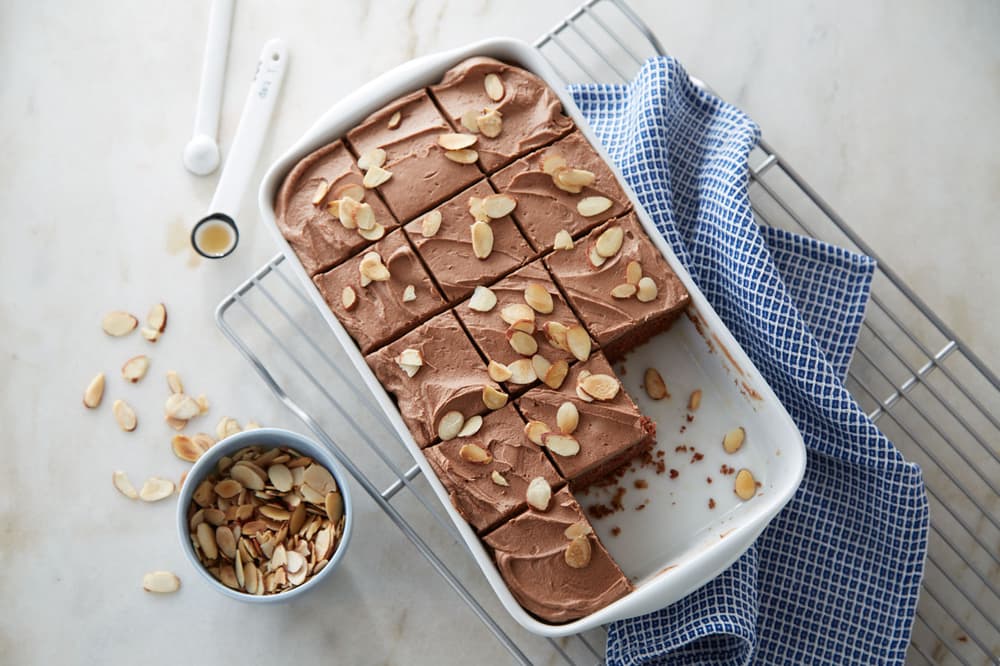 A rectangular dish of chocolate frosted cake topped with sliced almonds, partially cut into squares, sits on a wire rack next to a blue checkered towel and a bowl of almonds.