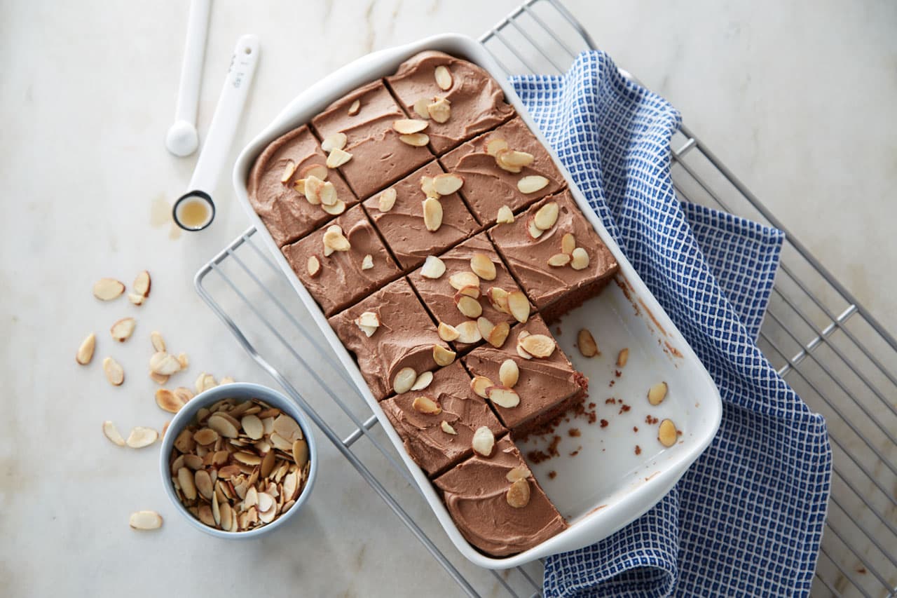 A rectangular dish of chocolate frosted cake topped with sliced almonds, partially cut into squares, sits on a wire rack next to a blue checkered towel and a bowl of almonds.