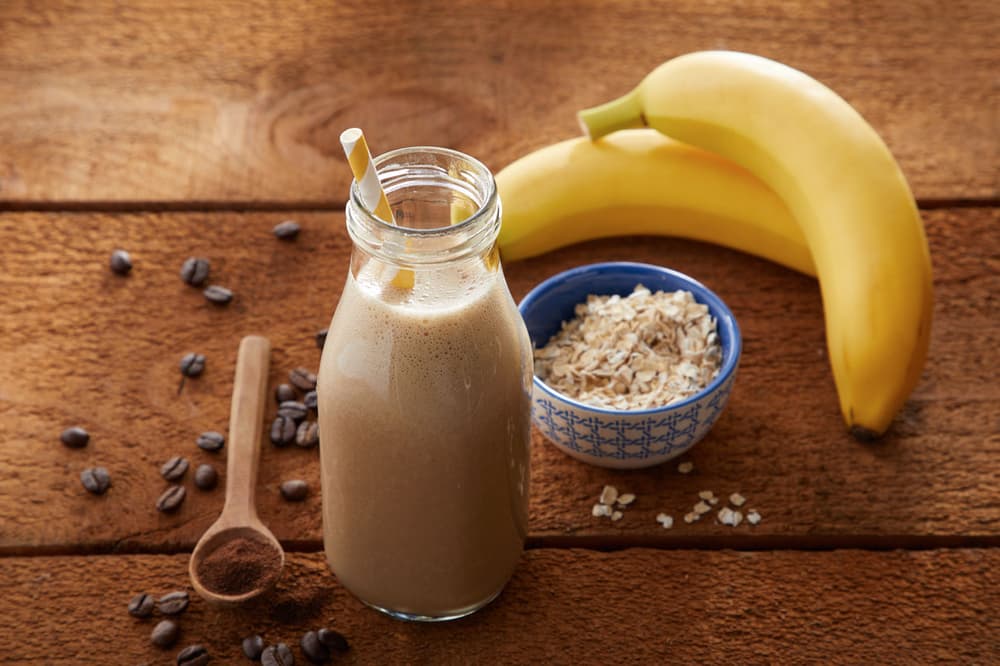 A glass bottle with a straw filled with a brown smoothie sits on a wooden surface beside two bananas, a bowl of oats, a wooden spoon with powder, and scattered coffee beans.
