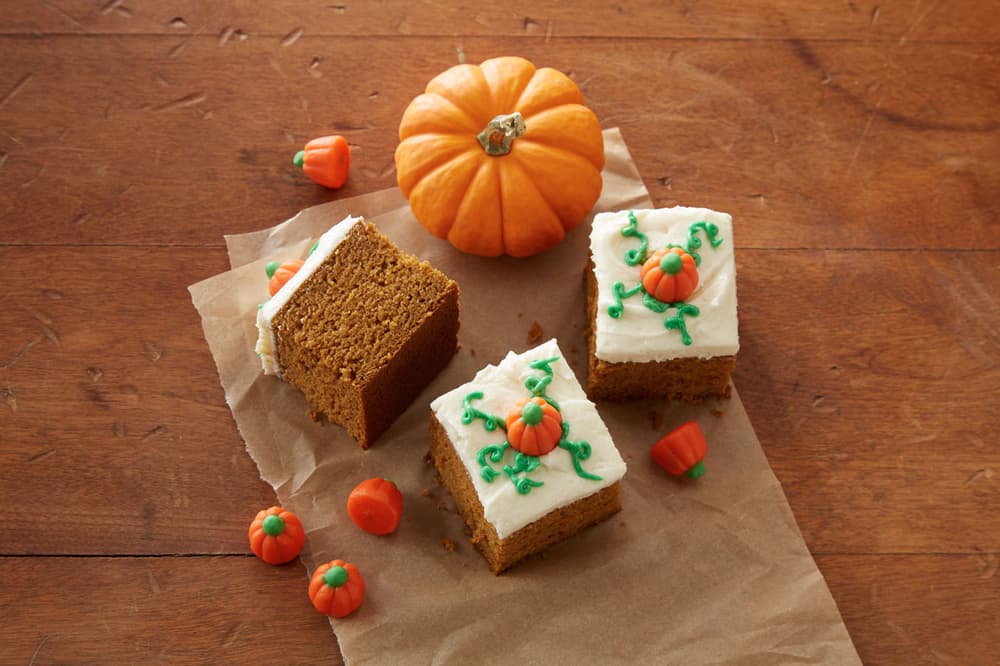 Three pumpkin bars with white frosting and pumpkin decorations, a small pumpkin, and candy pumpkins on a brown paper and wooden surface.