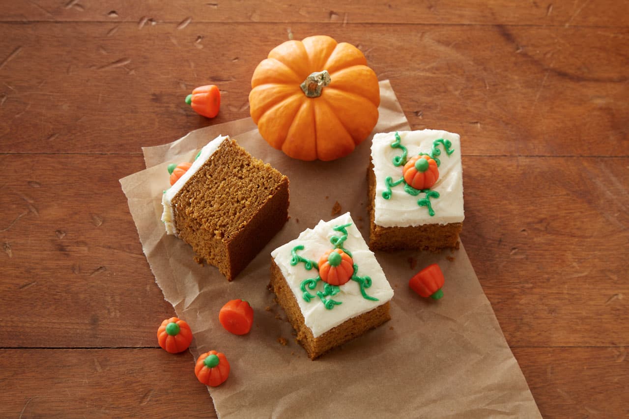 Three pumpkin bars with white frosting and pumpkin decorations, a small pumpkin, and candy pumpkins on a brown paper and wooden surface.