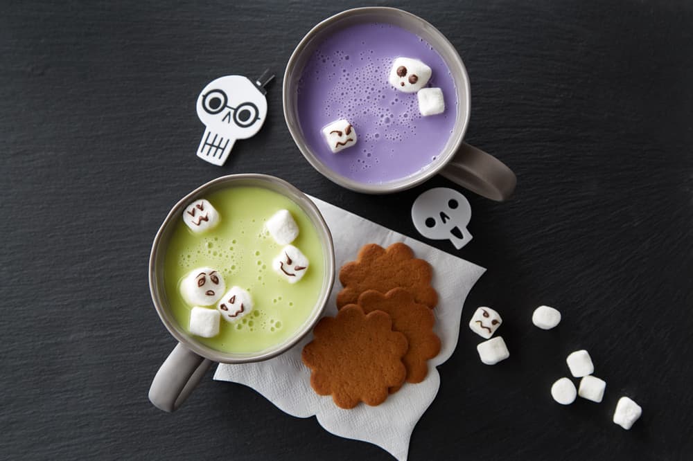 Two mugs with green and purple drinks, each topped with marshmallows decorated as faces, are on a black surface with ginger cookies and a few marshmallows, plus skull decorations.