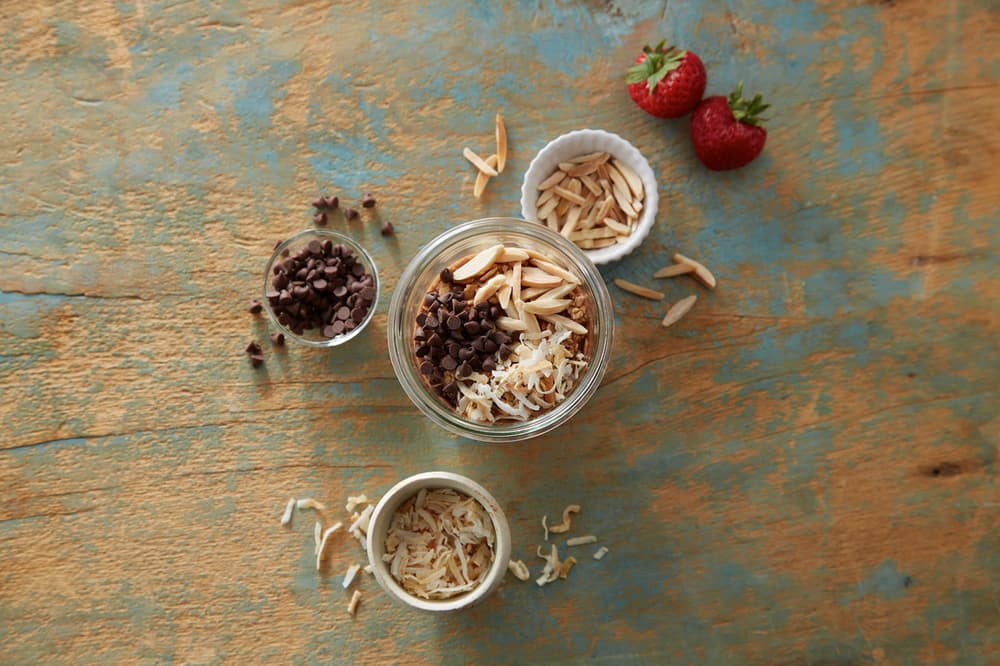 A glass jar filled with slivered almonds, chocolate chips, and shredded coconut is surrounded by small bowls of the same ingredients and two strawberries on a rustic blue wooden surface.