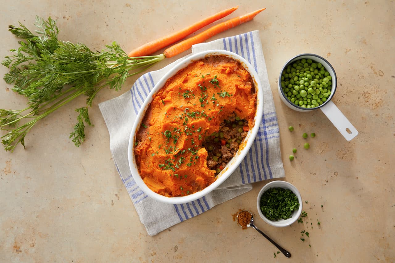 Oval baking dish with shepherds pie topped with mashed sweet potatoes, next to fresh carrots, peas in a bowl, chopped herbs, and a striped towel on a beige surface.