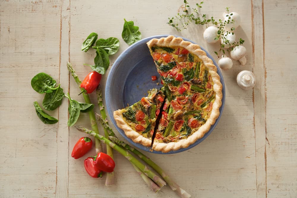 A vegetable quiche with a slice missing sits on a plate, surrounded by fresh spinach, red peppers, asparagus, mushrooms, and thyme on a light wooden table.