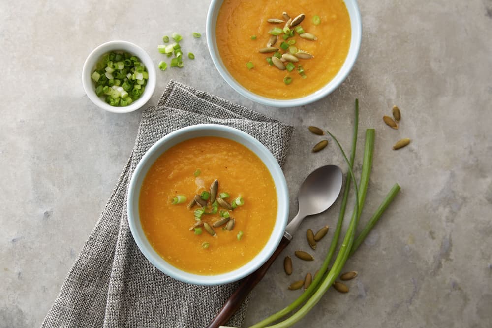 Two bowls of orange soup topped with green onions and seeds, next to a spoon, green onions, and a small bowl of chopped green onions on a gray surface.