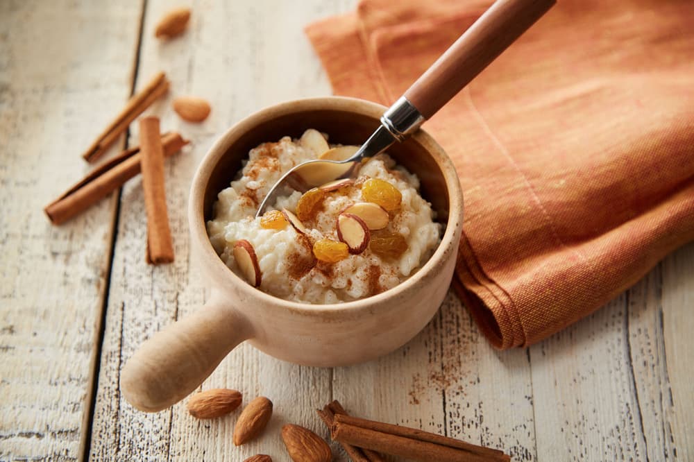 A wooden bowl of rice pudding topped with raisins and sliced almonds, with a spoon inside, sits on a rustic table next to cinnamon sticks, whole almonds, and an orange napkin.