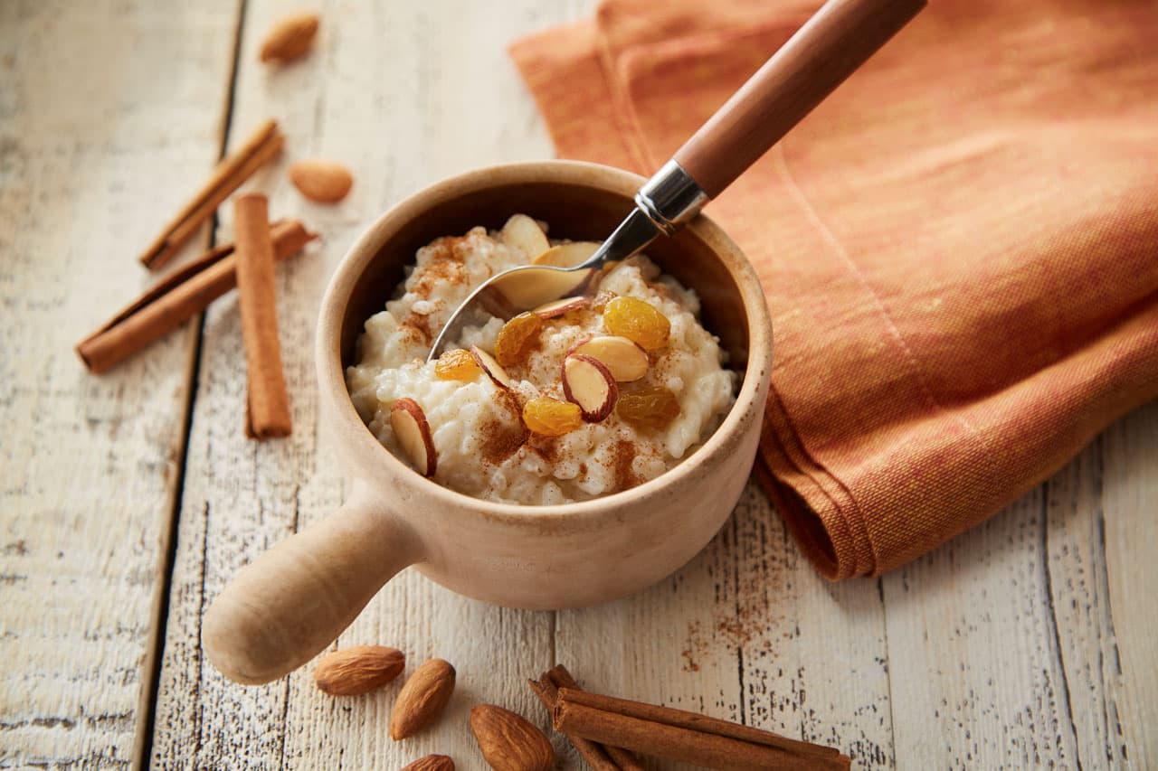 A wooden bowl of rice pudding topped with raisins and sliced almonds, with a spoon inside, sits on a rustic table next to cinnamon sticks, whole almonds, and an orange napkin.