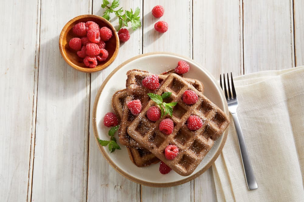 A plate of chocolate waffles topped with fresh raspberries and mint, next to a fork and a bowl of raspberries on a wooden table.