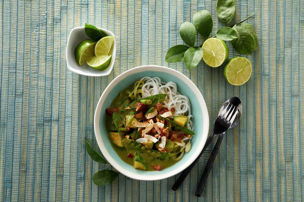 Bowl of noodles with green curry, vegetables, and herbs on a blue mat, with lime wedges and utensils beside the bowl.
