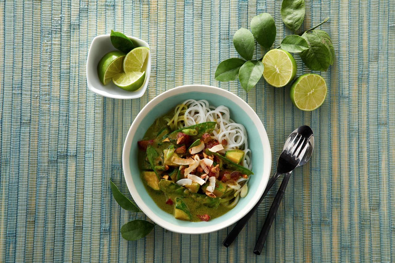Bowl of noodles with green curry, vegetables, and herbs on a blue mat, with lime wedges and utensils beside the bowl.