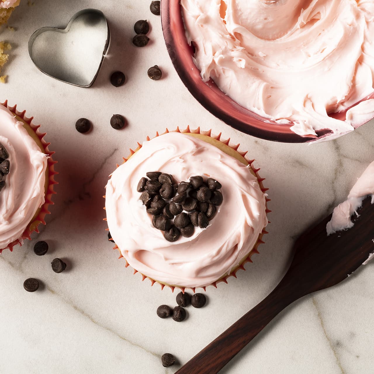 Cupcake with pink frosting and a heart shape made of chocolate chips on top, next to a bowl of frosting, spatula, heart-shaped cutter, and scattered chocolate chips on a marble surface.