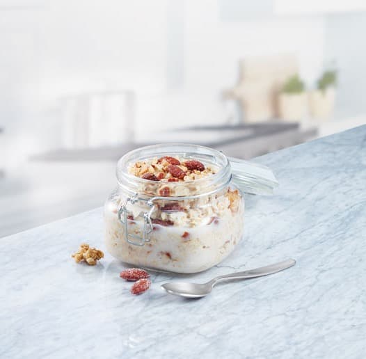 A glass jar filled with granola and milk sits on a marble countertop, with a spoon and a few granola pieces beside it.