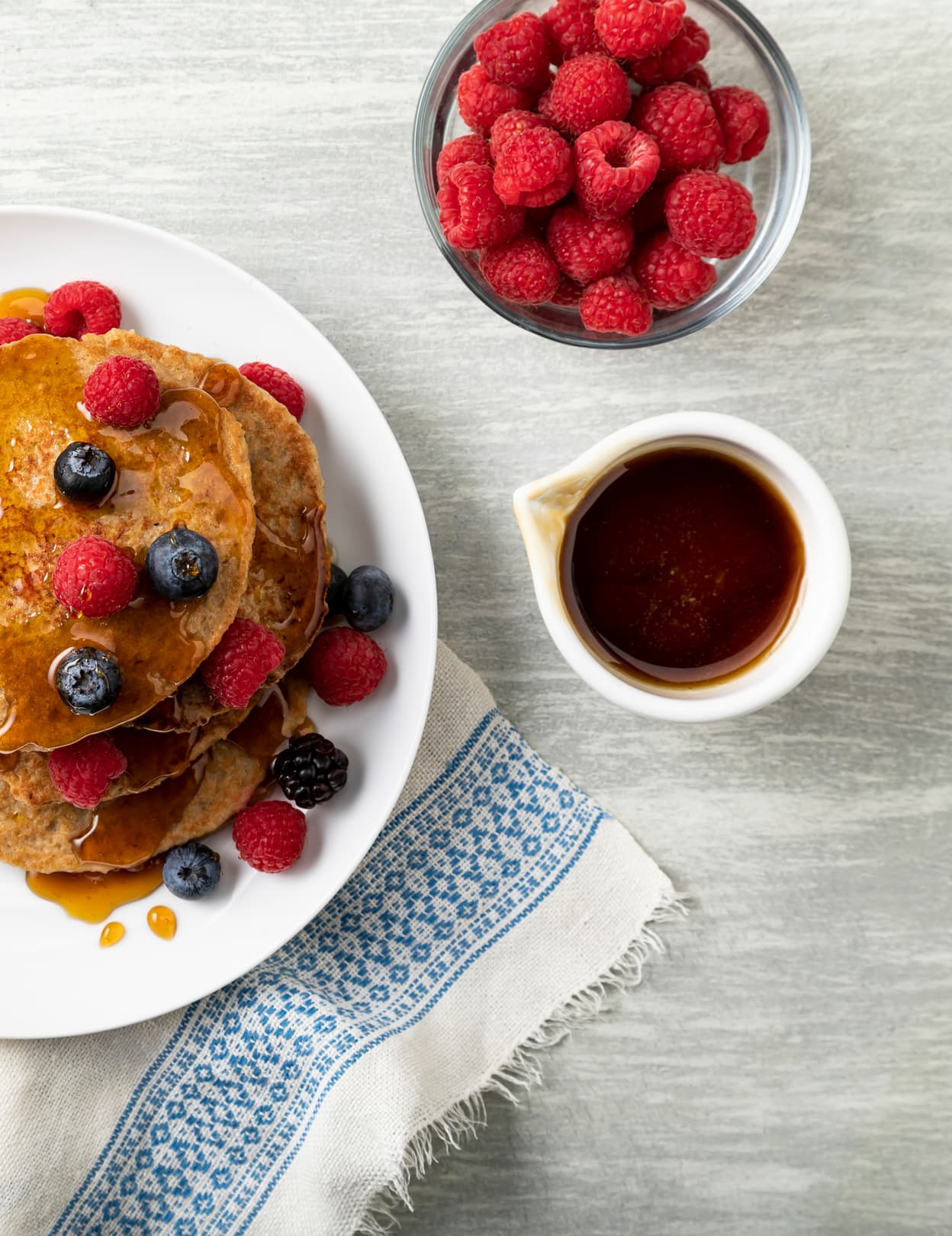 A plate of pancakes topped with berries and syrup, next to a bowl of raspberries and a small jug of syrup on a gray surface with a patterned cloth.