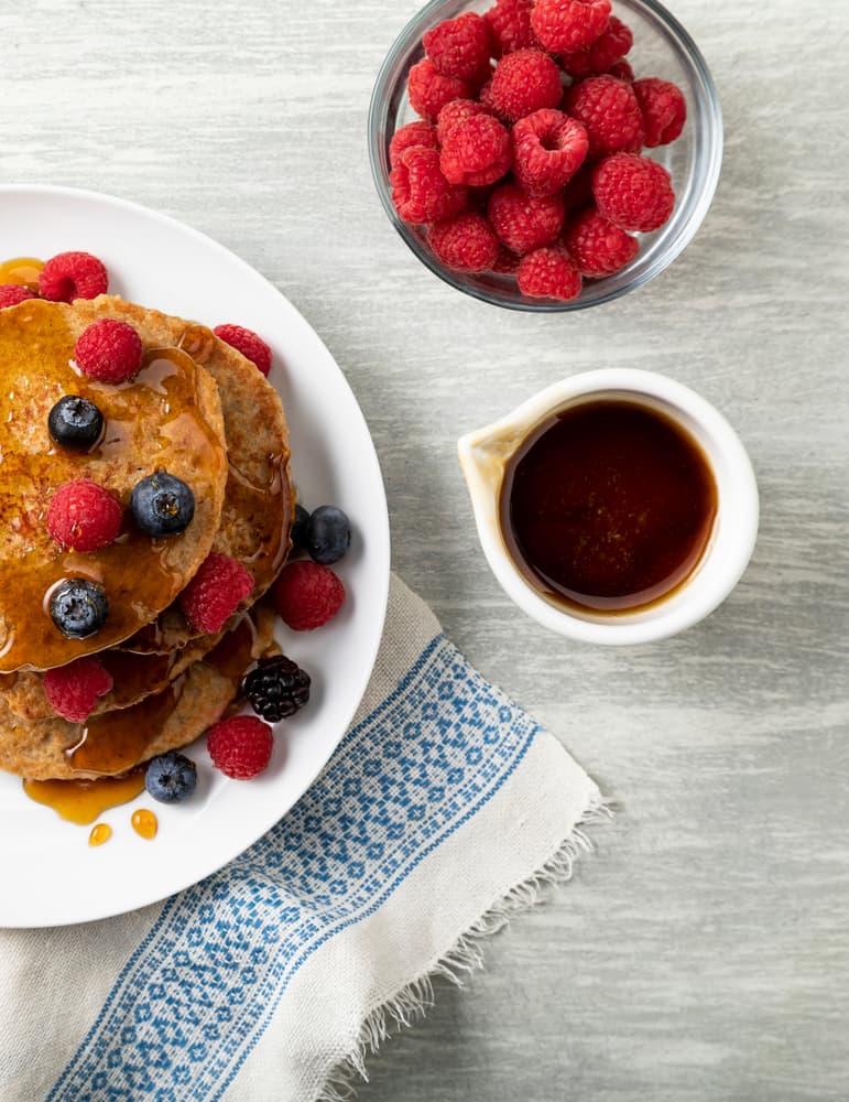 A plate of pancakes topped with berries and syrup, next to a bowl of raspberries and a small jug of syrup on a gray surface with a patterned cloth.