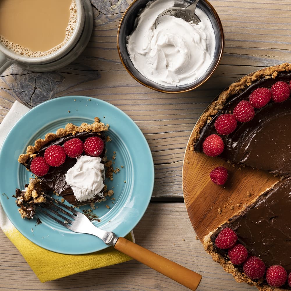 A slice of chocolate tart with raspberries and whipped cream on a blue plate, next to a cup of coffee and a bowl of whipped cream.