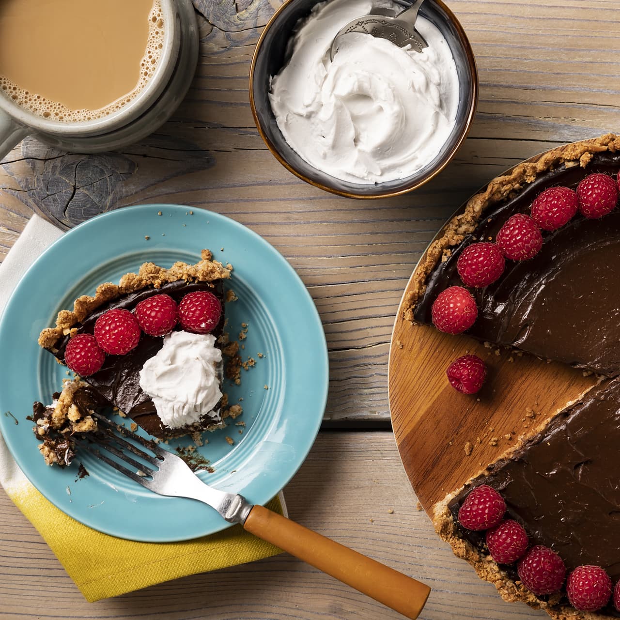 A slice of chocolate tart with raspberries and whipped cream on a blue plate, next to a cup of coffee and a bowl of whipped cream.