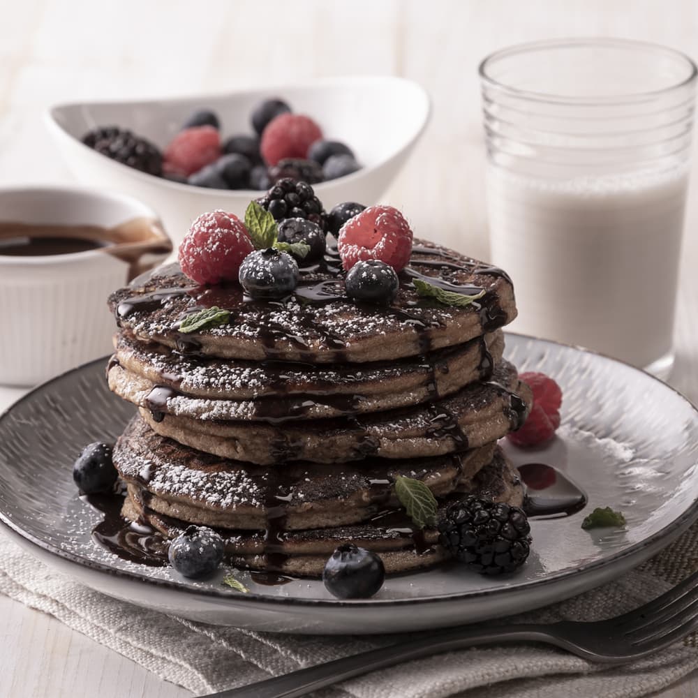 A stack of pancakes topped with berries, chocolate syrup, and powdered sugar, served with a glass of milk, syrup, and a bowl of mixed berries in the background.