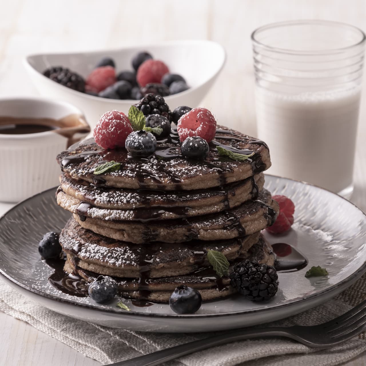 A stack of pancakes topped with berries, chocolate syrup, and powdered sugar, served with a glass of milk, syrup, and a bowl of mixed berries in the background.