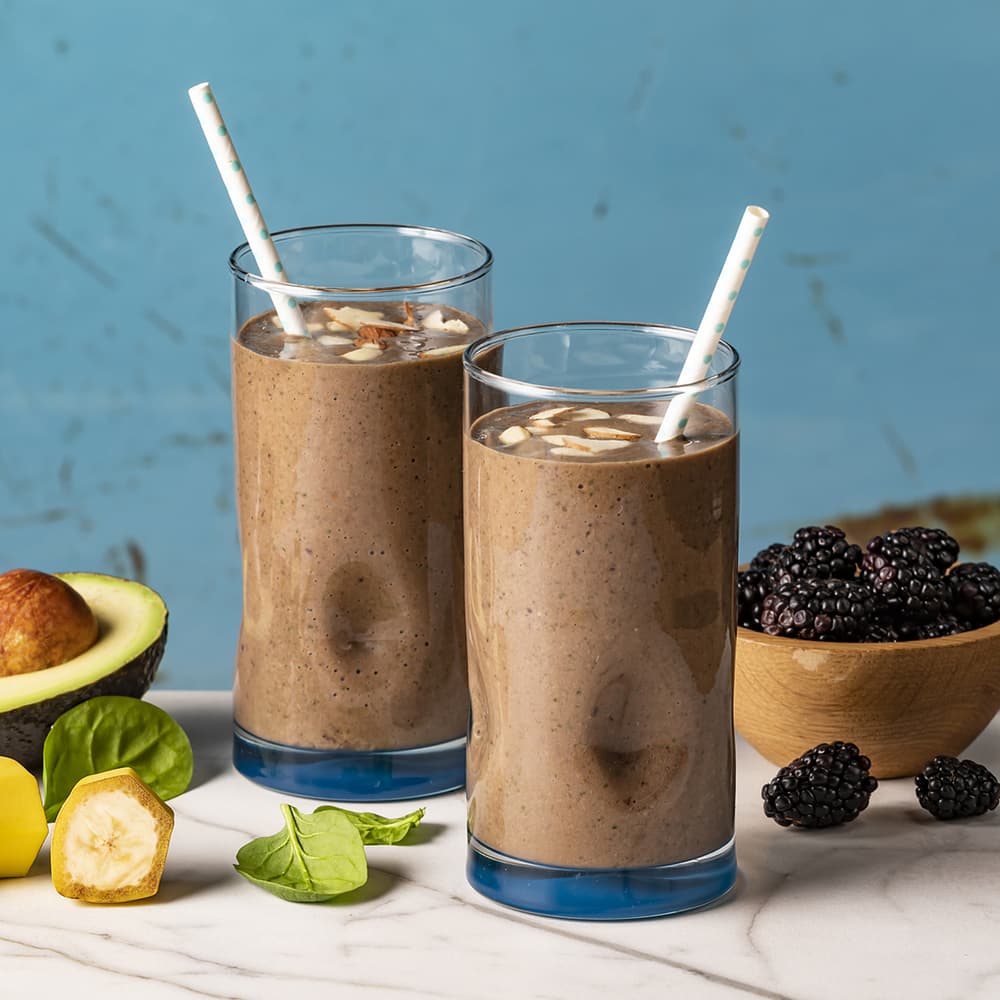 Two glasses of chocolate smoothie with almond slices and straws, surrounded by avocado, banana, spinach, and a wooden bowl of blackberries on a marble surface.
