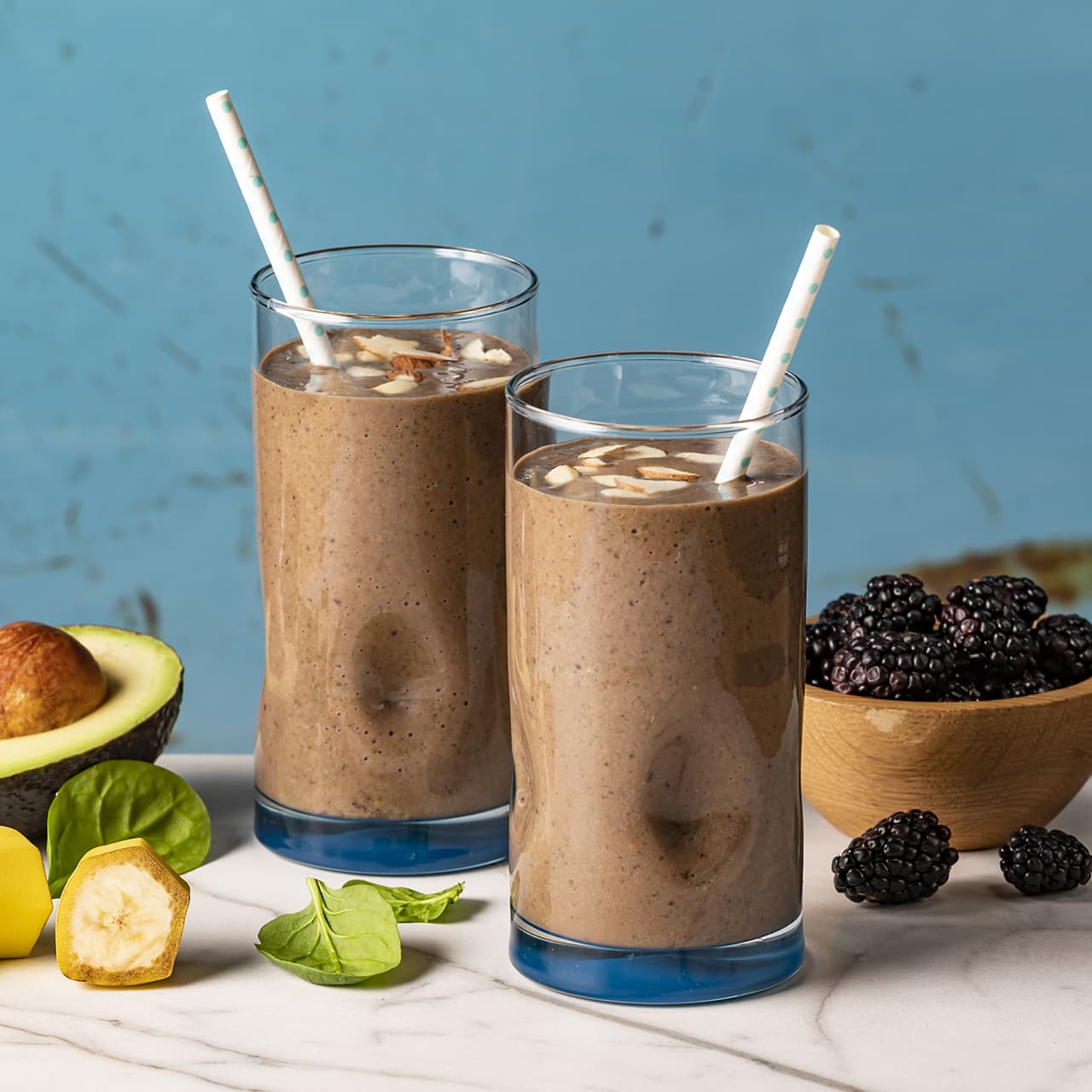 Two glasses of chocolate smoothie with almond slices and straws, surrounded by avocado, banana, spinach, and a wooden bowl of blackberries on a marble surface.