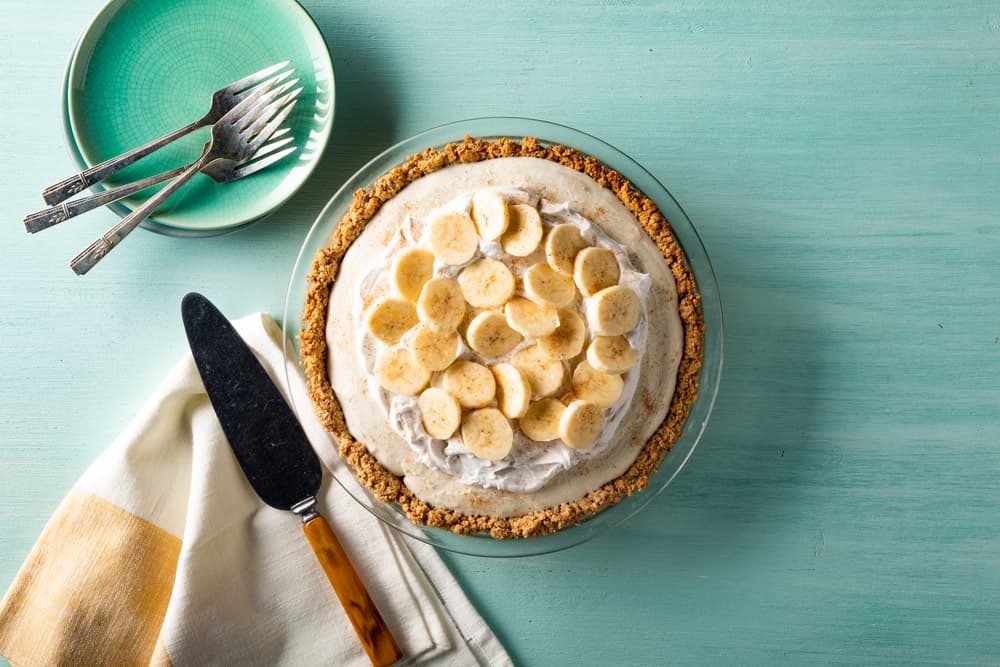 A banana cream pie with whipped topping and banana slices in a glass pie dish, next to a plate with forks, a pie server, and a folded napkin on a light turquoise surface.