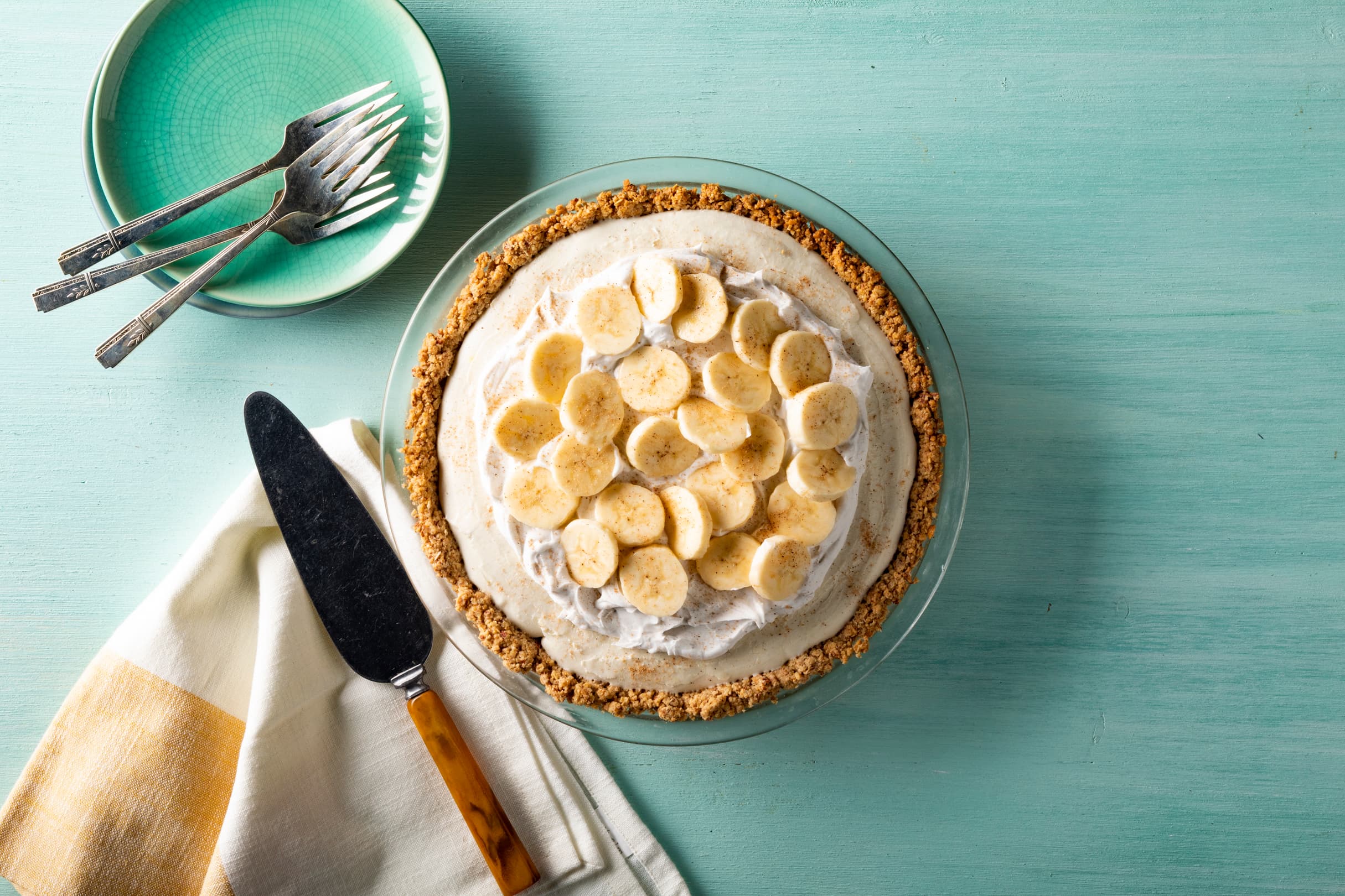 A banana cream pie with whipped topping and banana slices in a glass pie dish, next to a plate with forks, a pie server, and a folded napkin on a light turquoise surface.