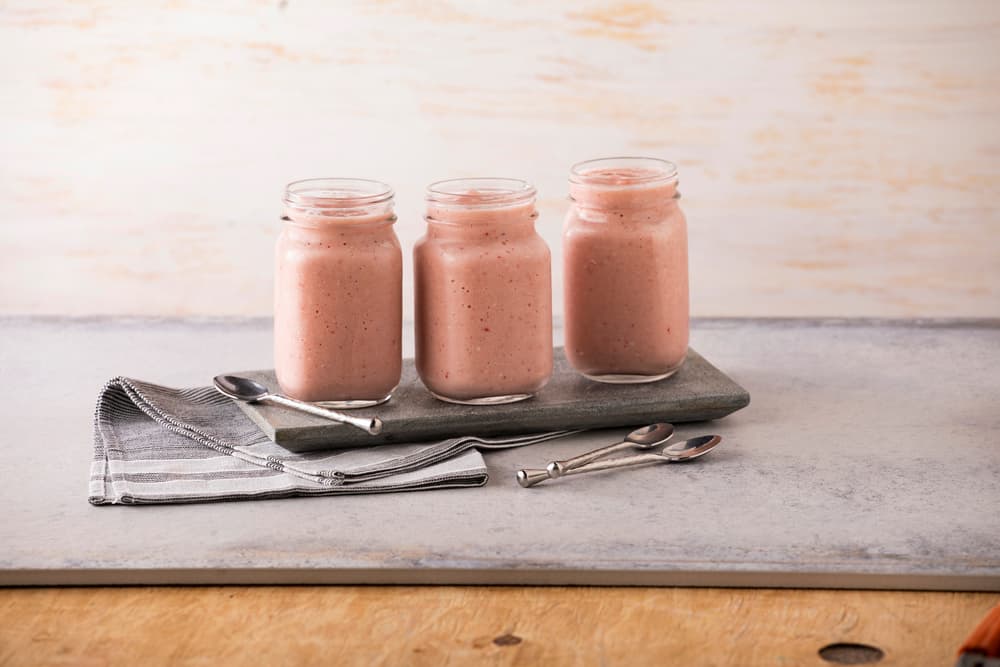 Three glass jars filled with pink smoothie are placed on a stone tray, with three spoons and a folded gray napkin beside them on a light surface.