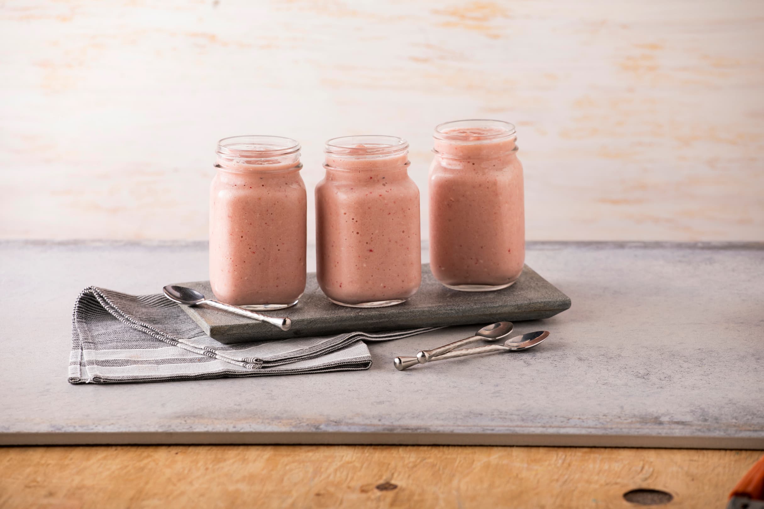 Three glass jars filled with pink smoothie are placed on a stone tray, with three spoons and a folded gray napkin beside them on a light surface.
