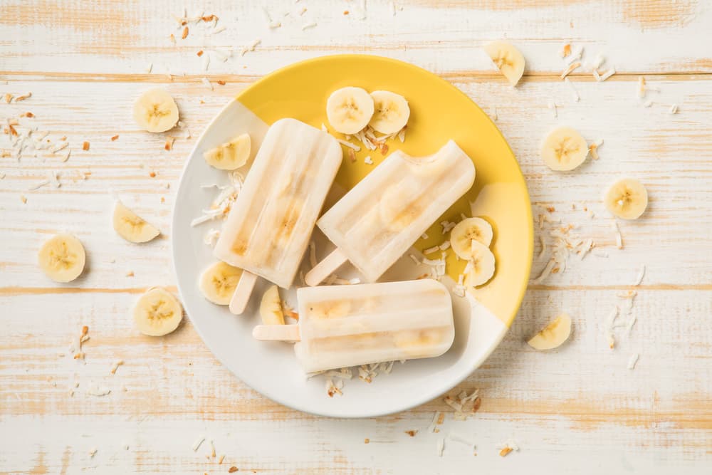 Three banana popsicles on a white and yellow plate, surrounded by banana slices and shredded coconut on a white wooden surface.