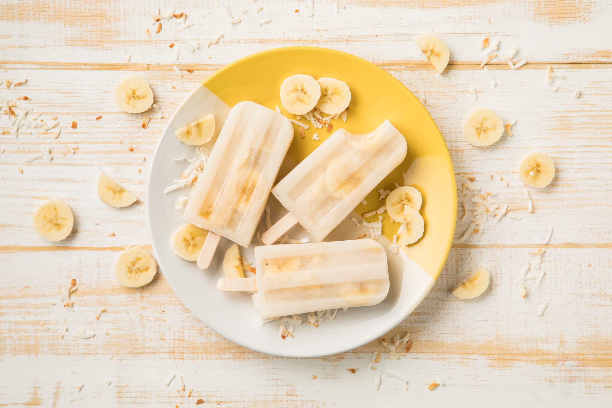 Three banana popsicles on a white and yellow plate, surrounded by banana slices and shredded coconut on a white wooden surface.