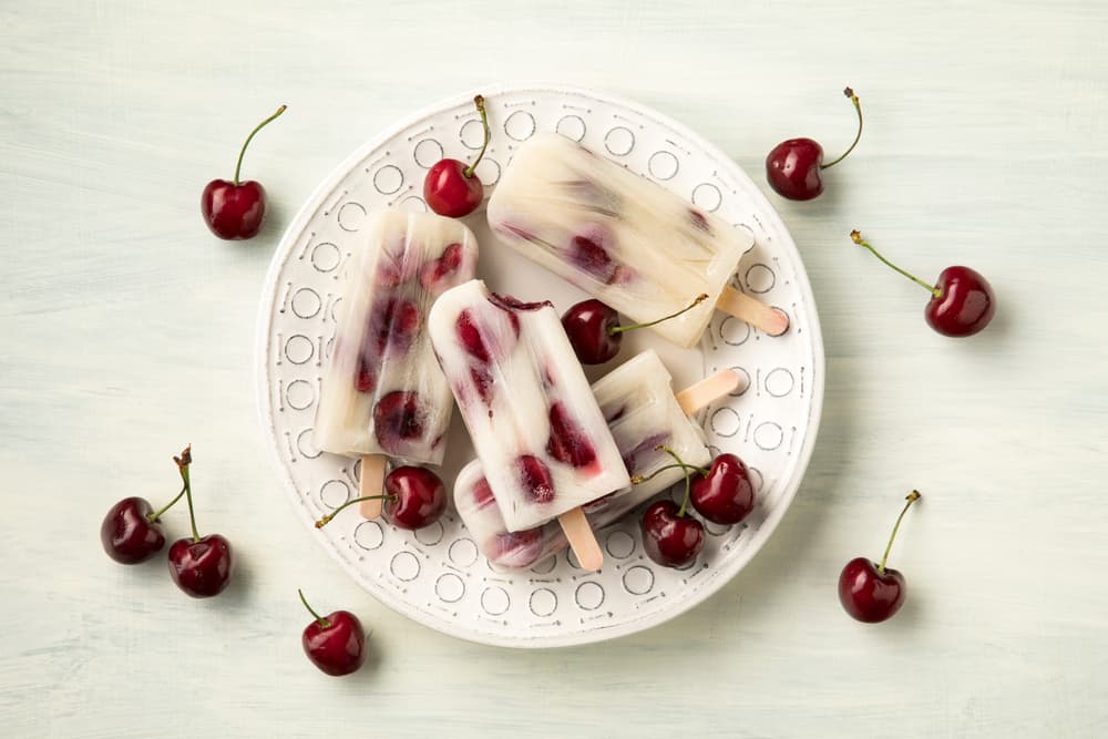 A white plate holds cherry popsicles and fresh cherries on a light background, with more cherries scattered around the plate.