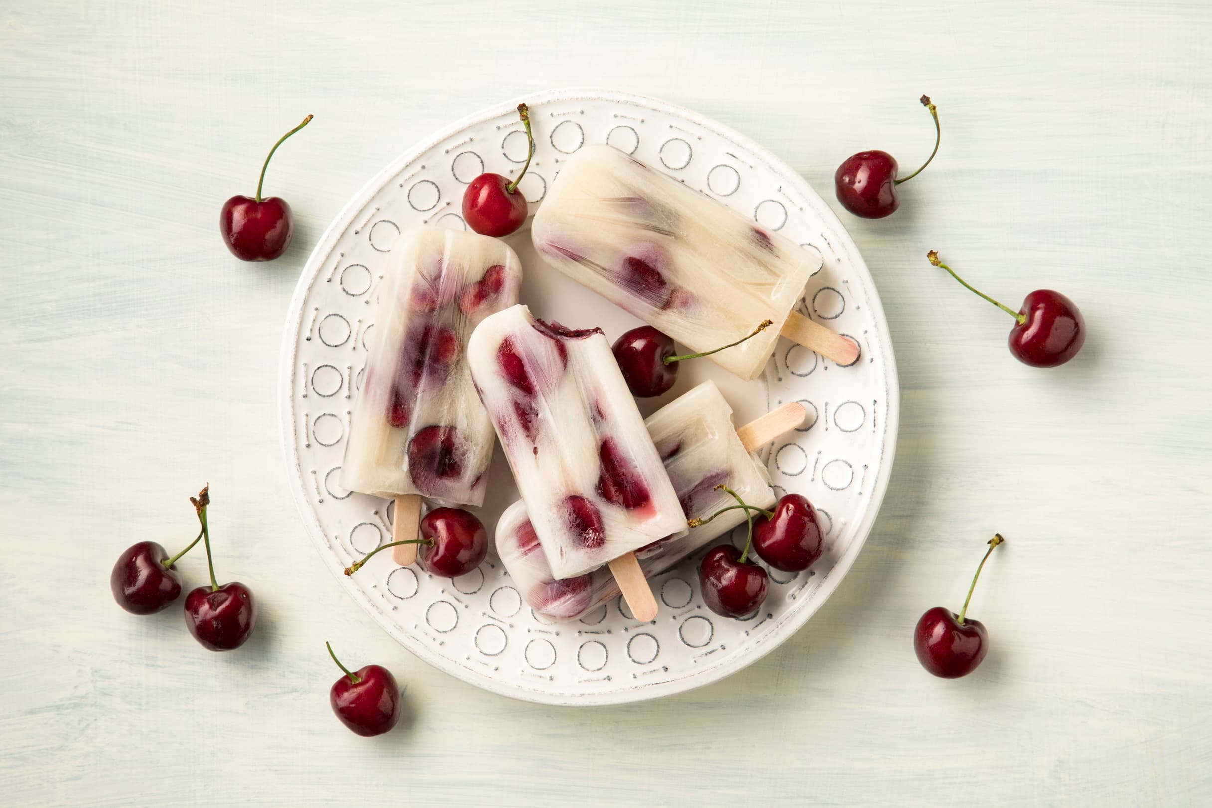 A white plate holds cherry popsicles and fresh cherries on a light background, with more cherries scattered around the plate.
