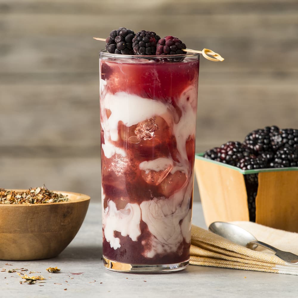 A tall glass of iced blackberry drink with swirled cream, garnished with fresh blackberries, sits on a table next to a bowl of blackberries and a bowl of dried tea leaves.