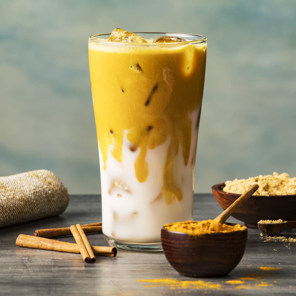 A tall glass of iced golden milk with turmeric, layered with milk and ice, sits on a table next to cinnamon sticks and a bowl of ground turmeric.