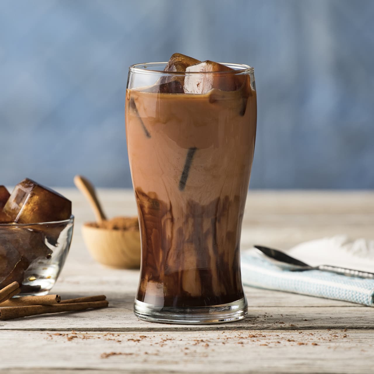 A glass of iced chocolate milk sits on a wooden table, with chocolate swirls, ice cubes, and cinnamon sticks nearby.