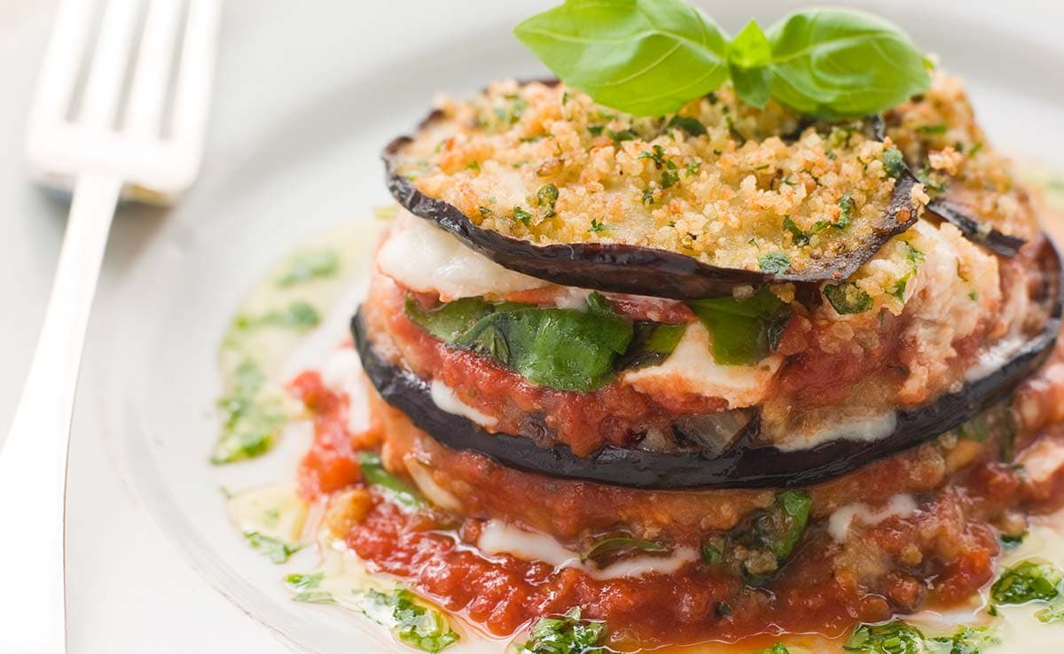 Stacked eggplant slices with tomato sauce, cheese, greens, and breadcrumbs, garnished with basil, served on a white plate with a fork.