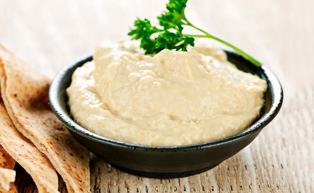 A black bowl filled with hummus, topped with a sprig of parsley, sits on a wooden surface next to pieces of flatbread.