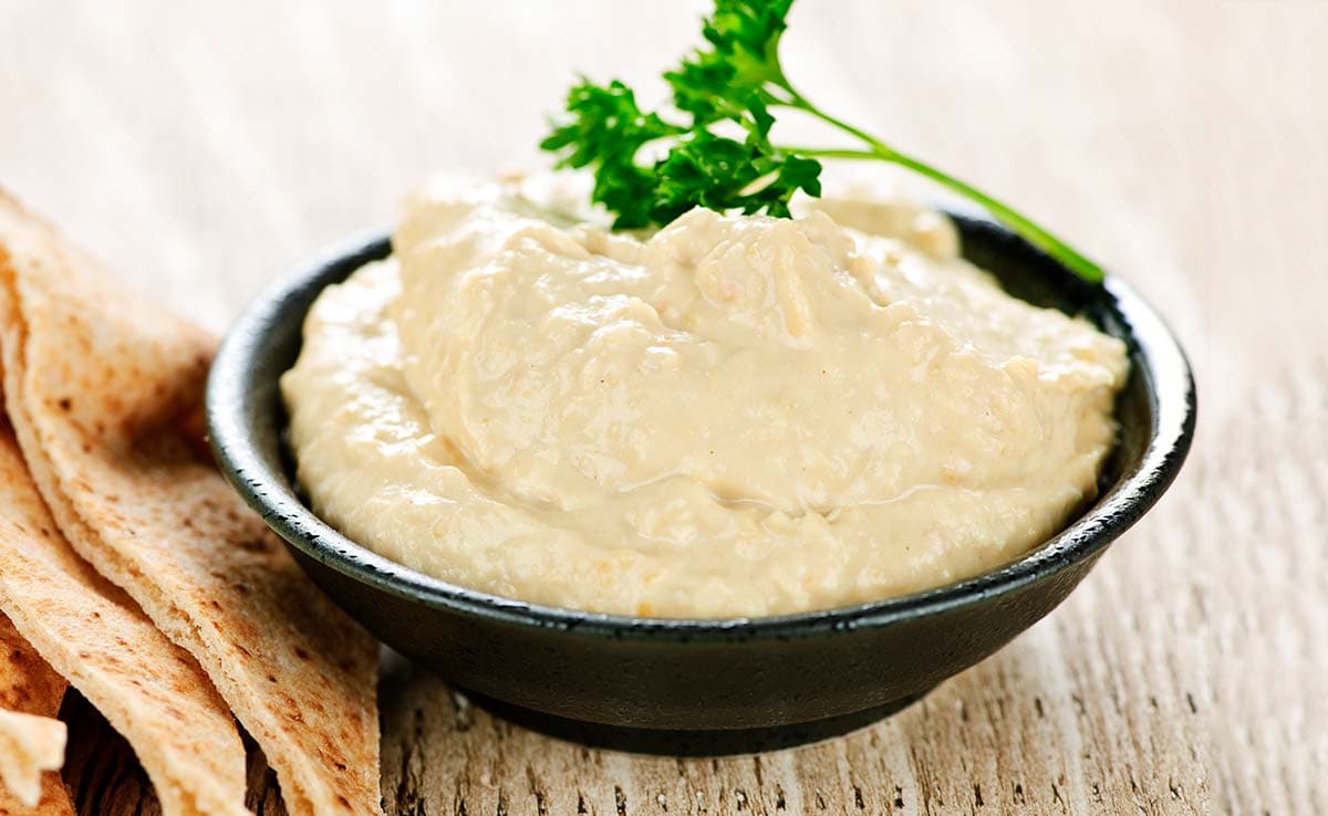 A black bowl filled with hummus, topped with a sprig of parsley, sits on a wooden surface next to pieces of flatbread.
