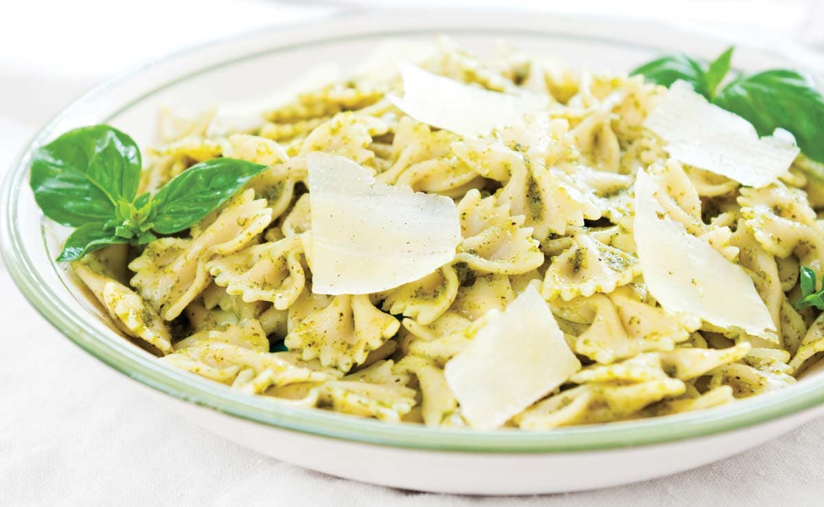 A bowl of farfalle pasta with green pesto sauce, garnished with fresh basil leaves and topped with shaved Parmesan cheese.